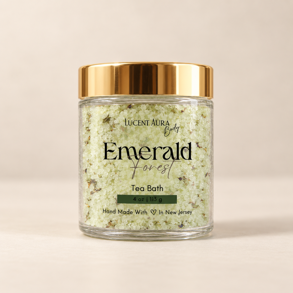 A clear glass jar of Lucent Aura Body Emerald Forest Tea Bath with a shiny gold lid, set against a warm beige background.
