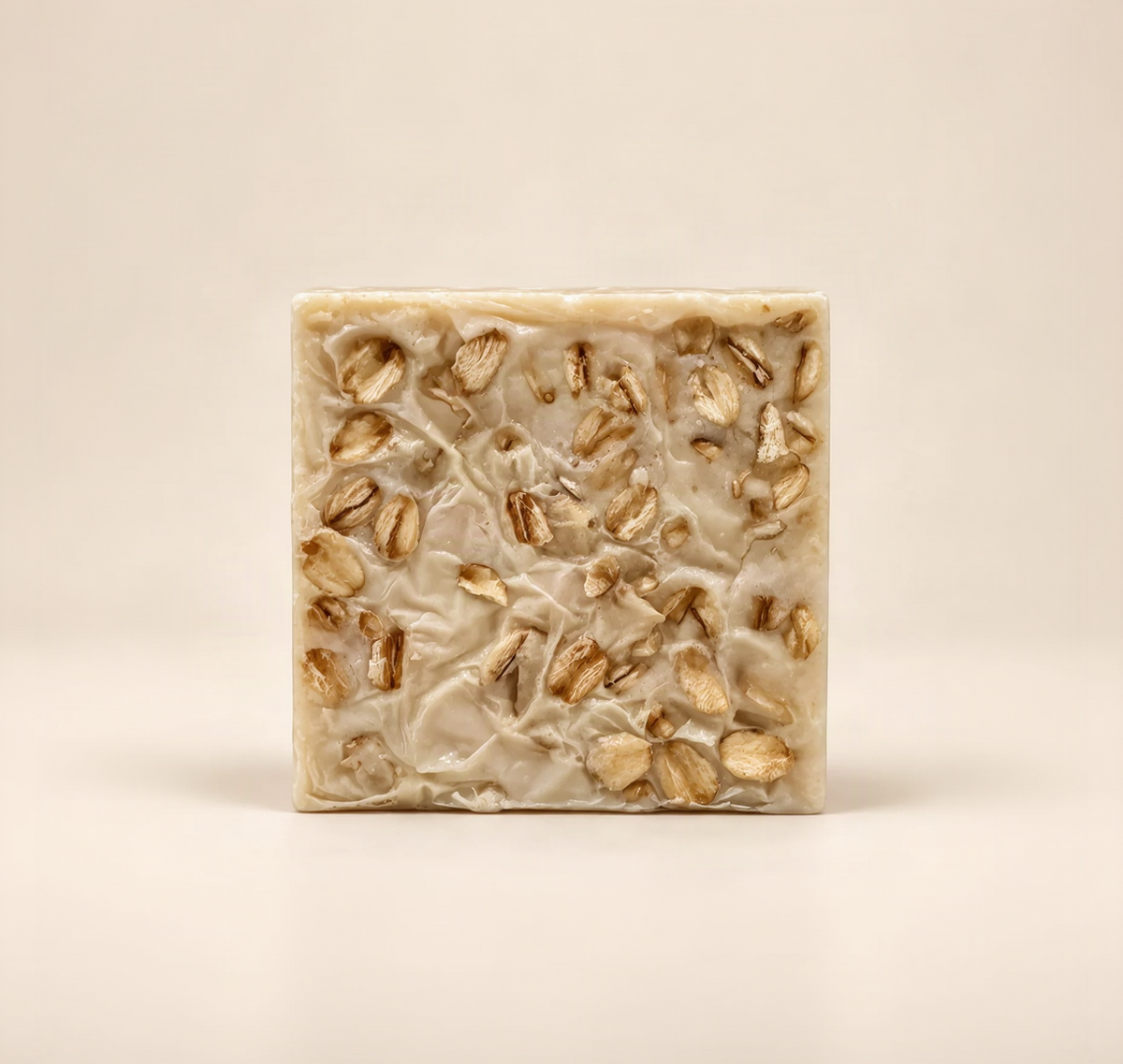 A square bar of handmade soap with a creamy beige base and scattered whole oats embedded throughout, photographed on a clean, solid, warm off-white background.