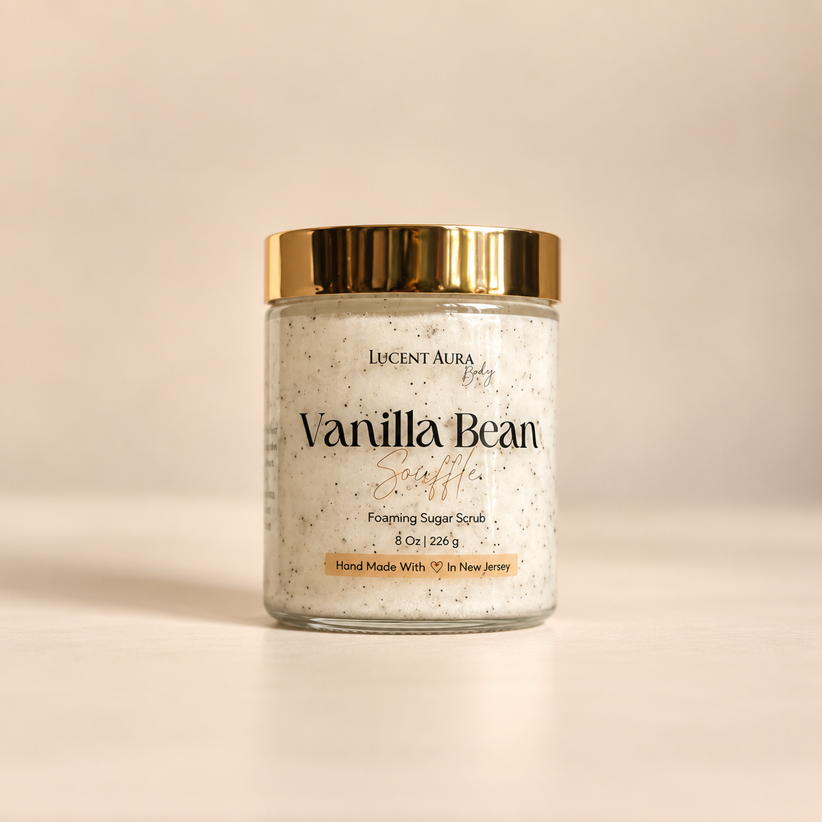 A jar of Lucent Aura Body Vanilla Bean Scuffie Foaming Sugar Scrub with a gold metallic lid, set against a warm beige background.