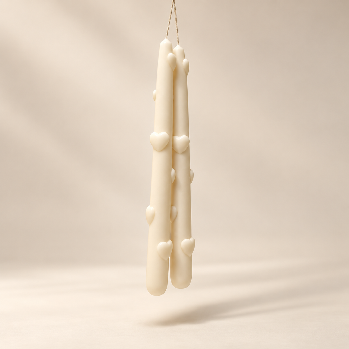 Two cream-colored taper candles with small raised heart details, hanging from a thin string against a warm beige studio background.