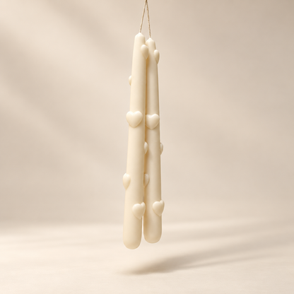 Two cream-colored taper candles with small raised heart details, hanging from a thin string against a warm beige studio background.