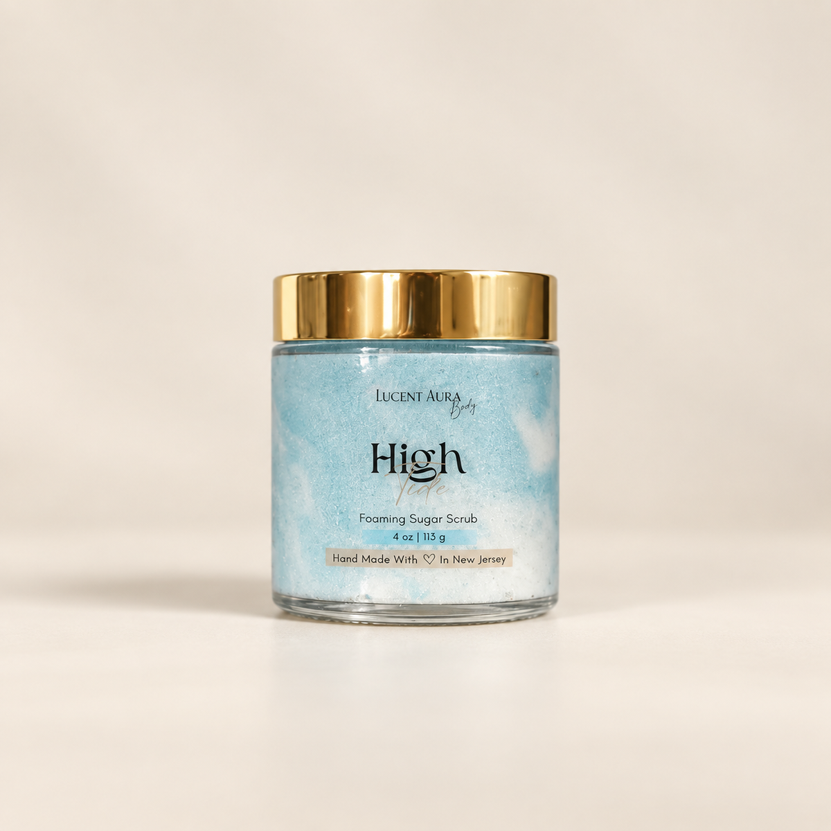 Lucint Aura High Tide Foaming Sugar Scrub in a clear glass jar with a gold lid and a light blue marbled label, set against a warm off-white background.