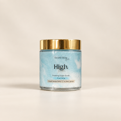 Lucint Aura High Tide Foaming Sugar Scrub in a clear glass jar with a gold lid and a light blue marbled label, set against a warm off-white background.