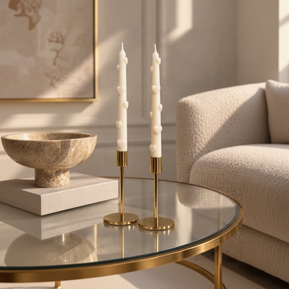 Two white taper candles with sculpted heart details on their wicks, displayed in elegant gold metal candlesticks on a round glass-top table in a softly lit living room.