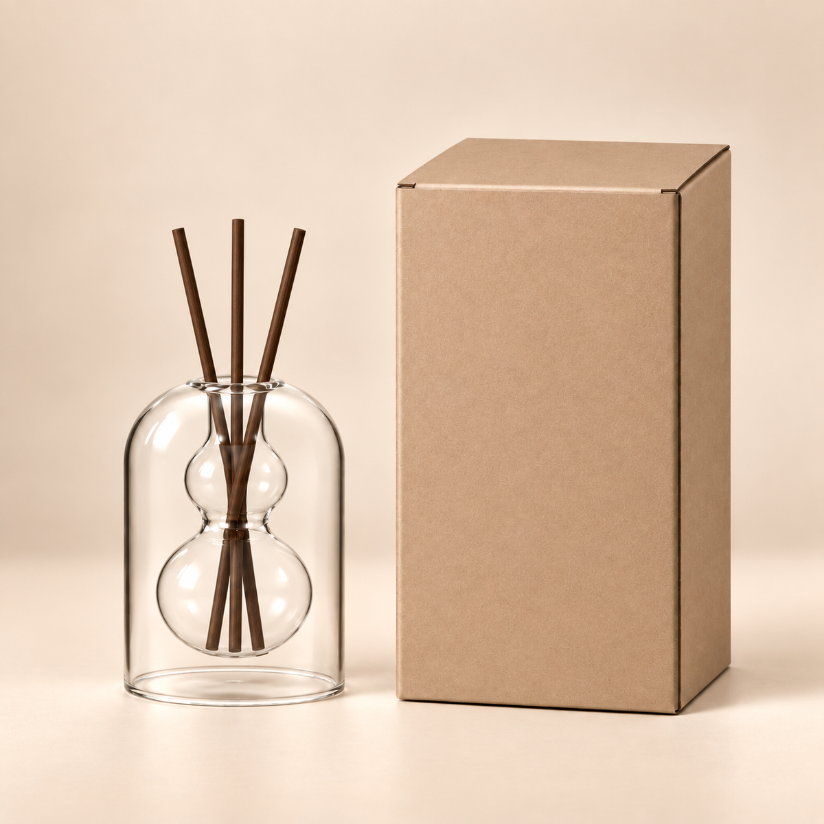 A clear glass reed diffuser with two bulb-shaped chambers and four dark brown reeds, standing beside a plain kraft paper box on a warm beige studio background.