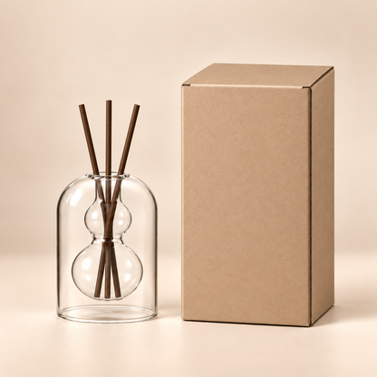 A clear glass reed diffuser with two bulb-shaped chambers and four dark brown reeds, standing beside a plain kraft paper box on a warm beige studio background.