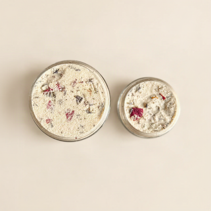 Two open clear jars filled with a light beige textured buttermilk powder, one large and one small, with visible pink and dark red rose petals, photographed on a plain warm beige background.