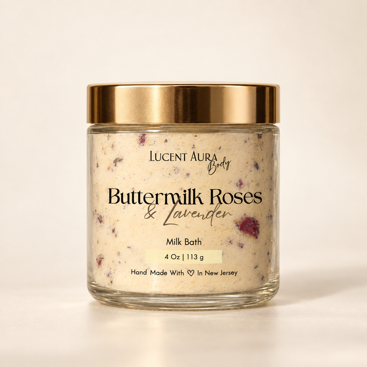 A glass jar of Lucent Aura Buttermilk Roses & Lavender Milk Bath with a gold metallic lid, set against a clean, warm off-white background.
