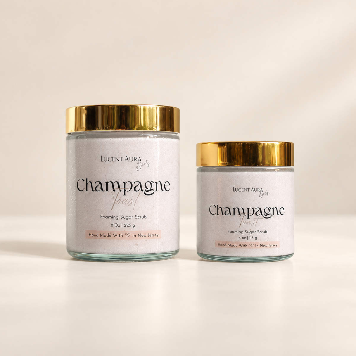 Two glass jars of Lucent Aura Champagne Toast Foaming Sugar Scrub with gold lids, one larger and one smaller, on a clean white background.