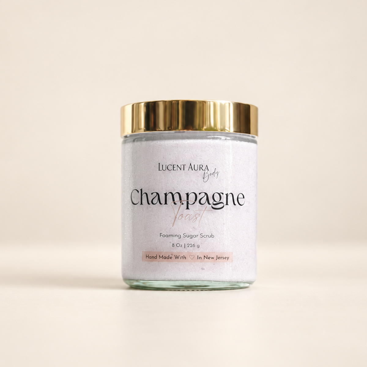A glass jar of Lucent Aura Champagne Toast Foaming Sugar Scrub with a shiny gold lid and a white label, set against a warm beige background.