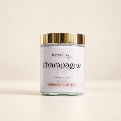A glass jar of Lucent Aura Champagne Toast Foaming Sugar Scrub with a shiny gold lid and a white label, set against a warm beige background.