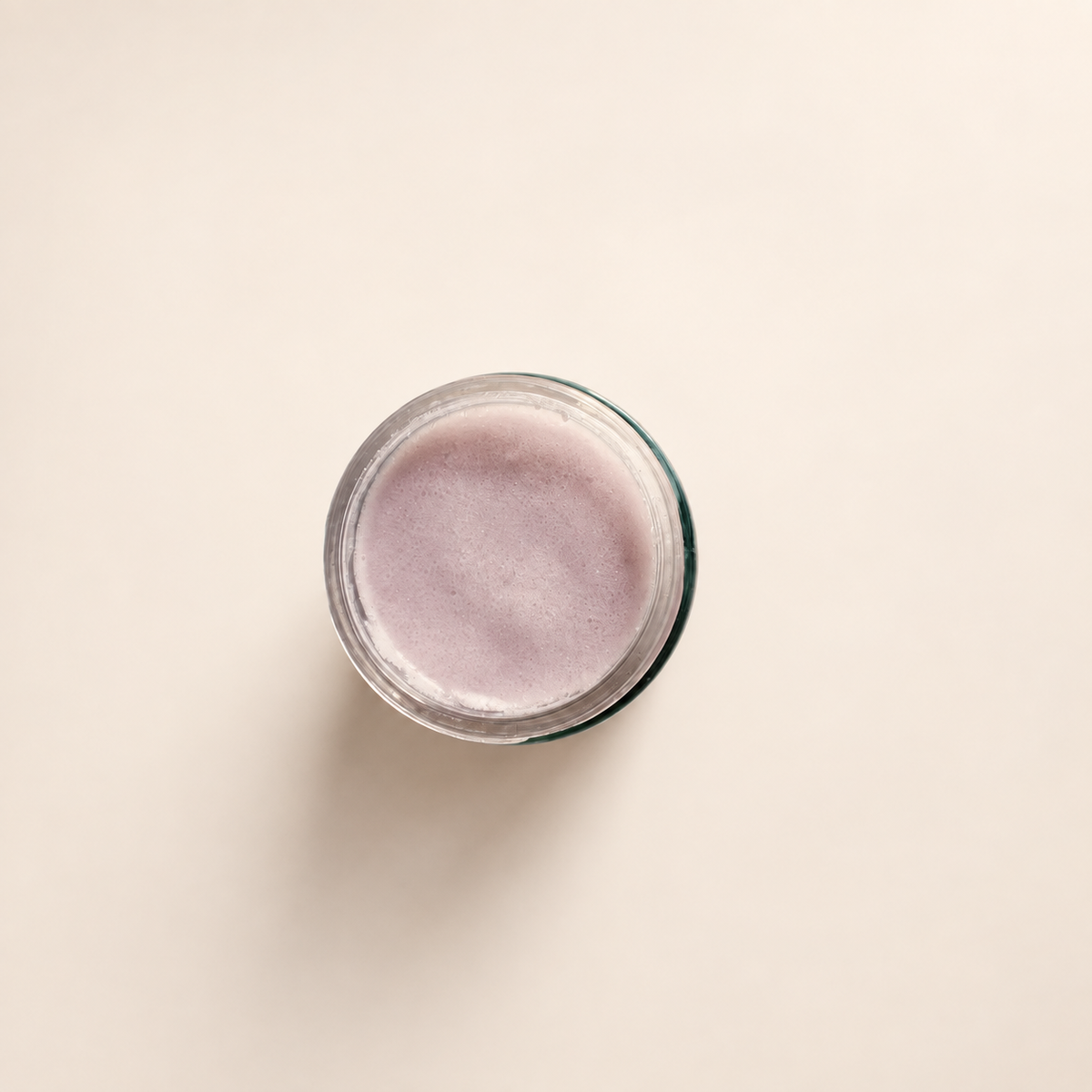 A top-down view of a clear glass jar containing a light lavender-purple sugar scrub, set against a solid off-white background.