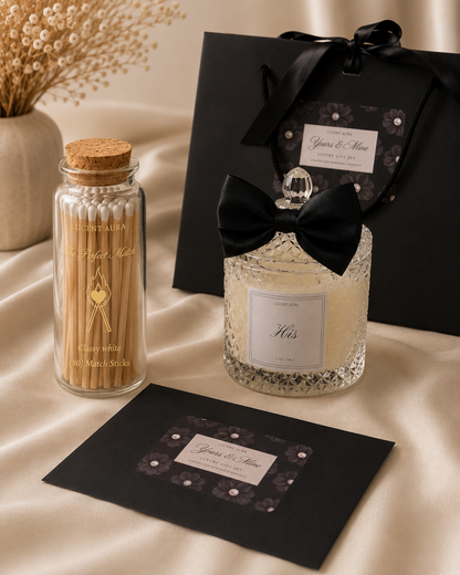 A luxury gift set featuring a clear glass jar candle with a black ribbon bow and silver glitter, a glass jar of wooden toothpicks, and a black gift box and card set, all arranged on a beige satin fabric background.