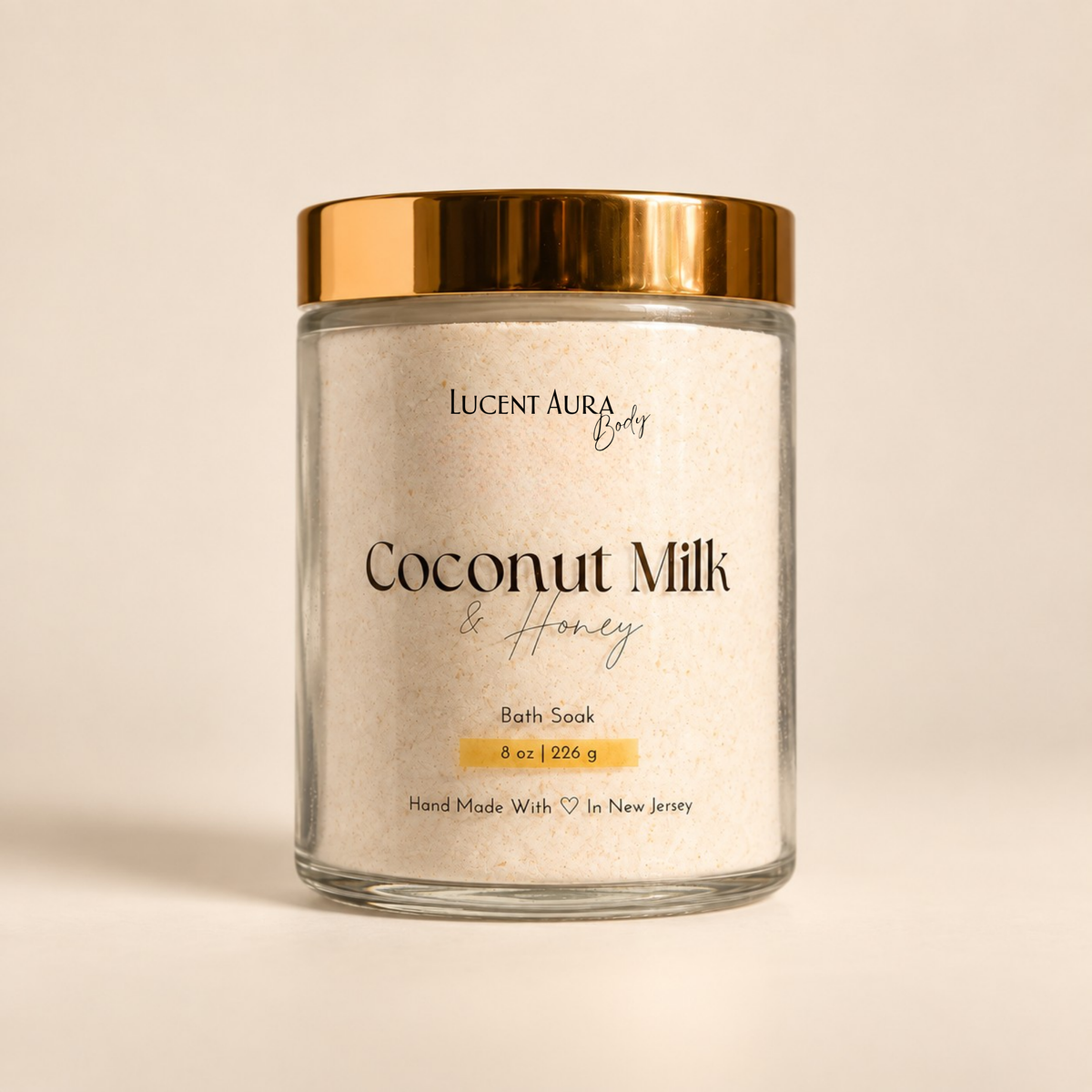 A glass jar of Lucent Aura Coconut Milk & Honey Bath Soak with a metallic gold lid, set against a warm beige background.