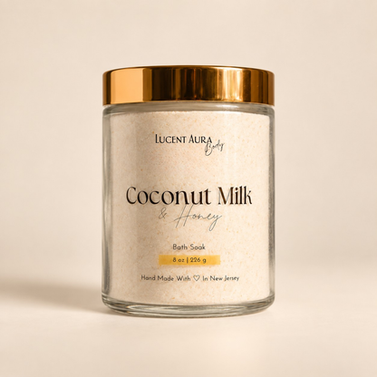 A glass jar of Lucent Aura Coconut Milk & Honey Bath Soak with a metallic gold lid, set against a warm beige background.