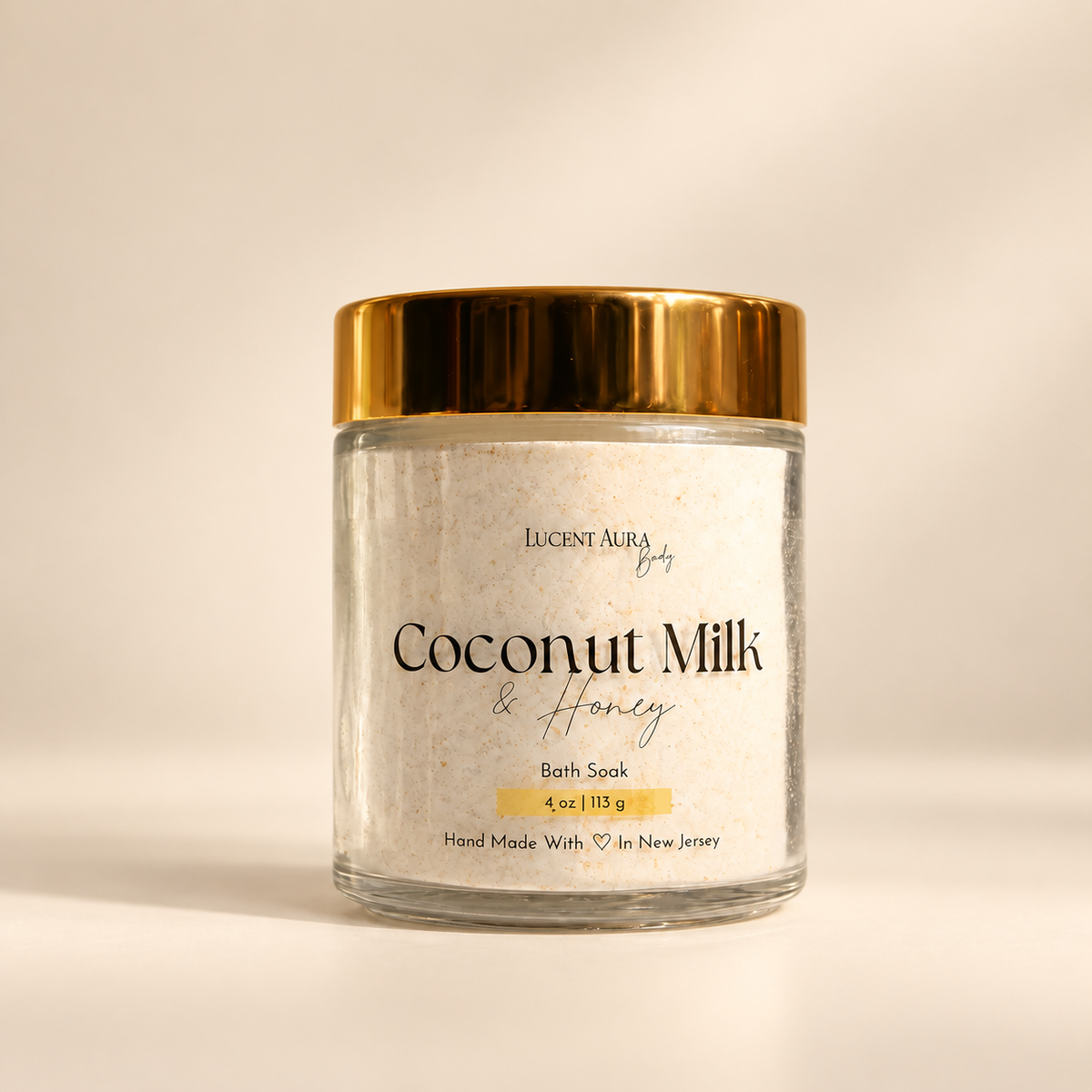 A clear glass jar of Lucent Aura Coconut Milk & Honey Bath Soak, featuring a shiny gold metallic lid and a white label, set against a warm beige background.