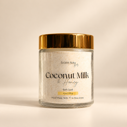 A clear glass jar of Lucent Aura Coconut Milk & Honey Bath Soak, featuring a shiny gold metallic lid and a white label, set against a warm beige background.