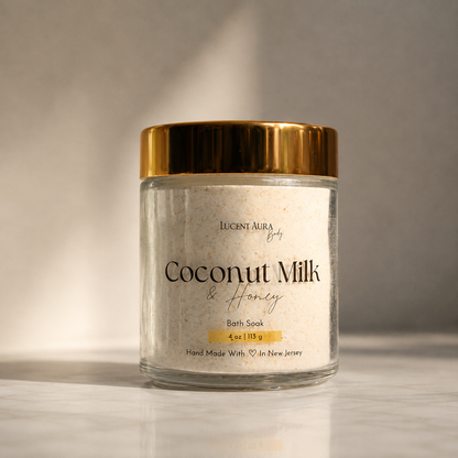 A glass jar of Lucent Aura Coconut Milk & Honey Bath Soak, featuring a metallic gold lid and a white label, set on a light marble surface with a neutral gray background.