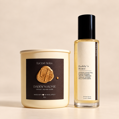 A candle jar and a fragrance oil bottle from the brand Lucent Aura, both labeled 'Daddy's Home', displayed on a warm beige background. The candle is cream-colored in a cream-colored jar with a dark brown label featuring a gold wax seal.