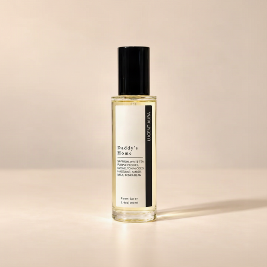 A clear glass bottle of room spray with a black cap and a white label reading 'Daddy's Home' and 'LUCENT AURA', filled with pale yellow liquid, set against a warm beige background.