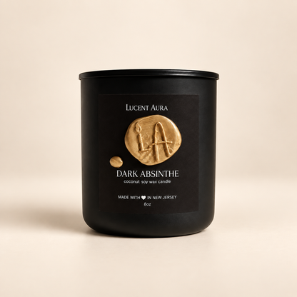 A matte black candle jar with a black label featuring a gold wax seal and the brand name 'Lucent Aura' and scent 'Dark Absinthe', set against a warm beige background.