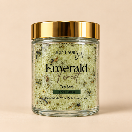 A clear glass jar of Lucent Aura Body Emerald Forest Tea Bath with a shiny gold lid, set against a solid beige background.