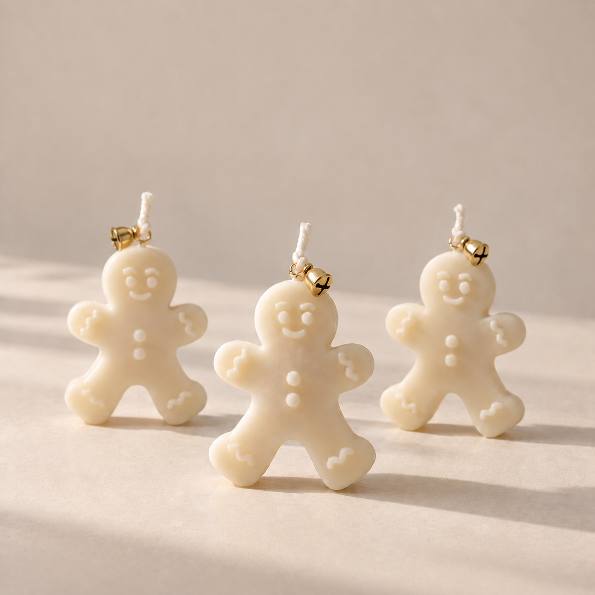Three small Gingerbread man candles in an off-white/cream color with gold-toned bells, arranged on a warm beige surface against a matching neutral background.