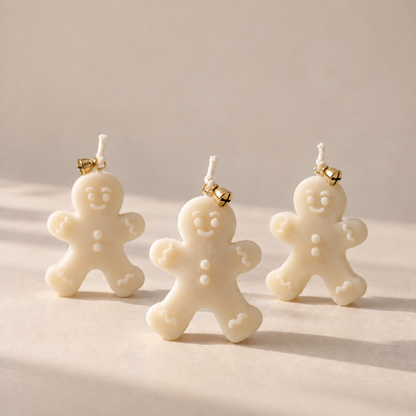 Three small Gingerbread man candles in an off-white/cream color with gold-toned bells, arranged on a warm beige surface against a matching neutral background.