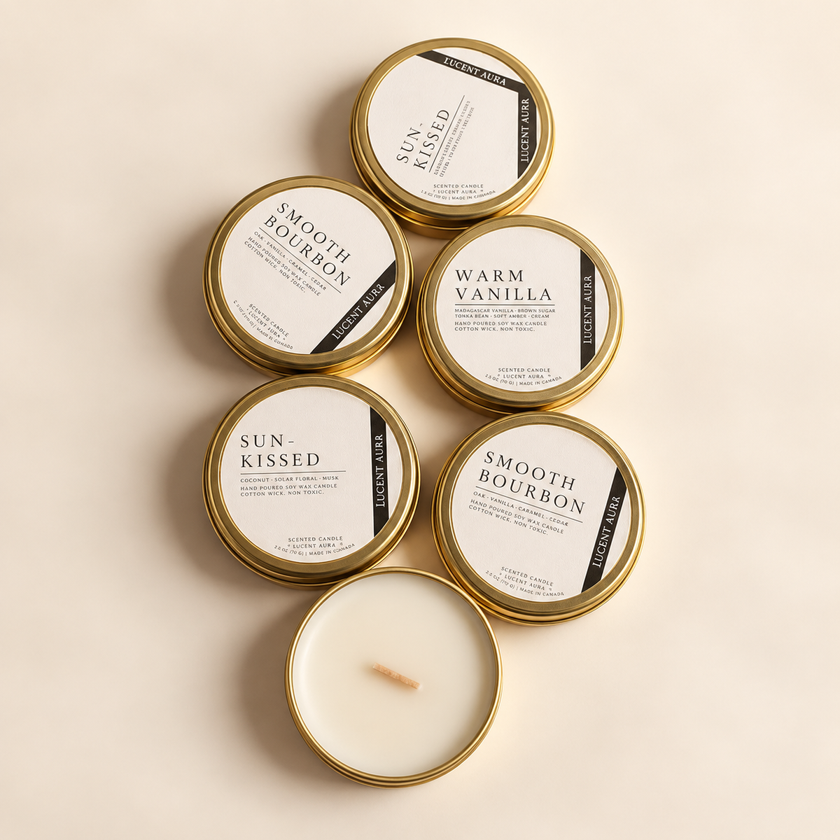 Six soy candles in round gold metal tins arranged on a warm cream background. Five tins are closed with white labels reading Smooth Bourbon, Warm Vanilla, and Sun Kissed, and one tin is open showing the white wax and wick.