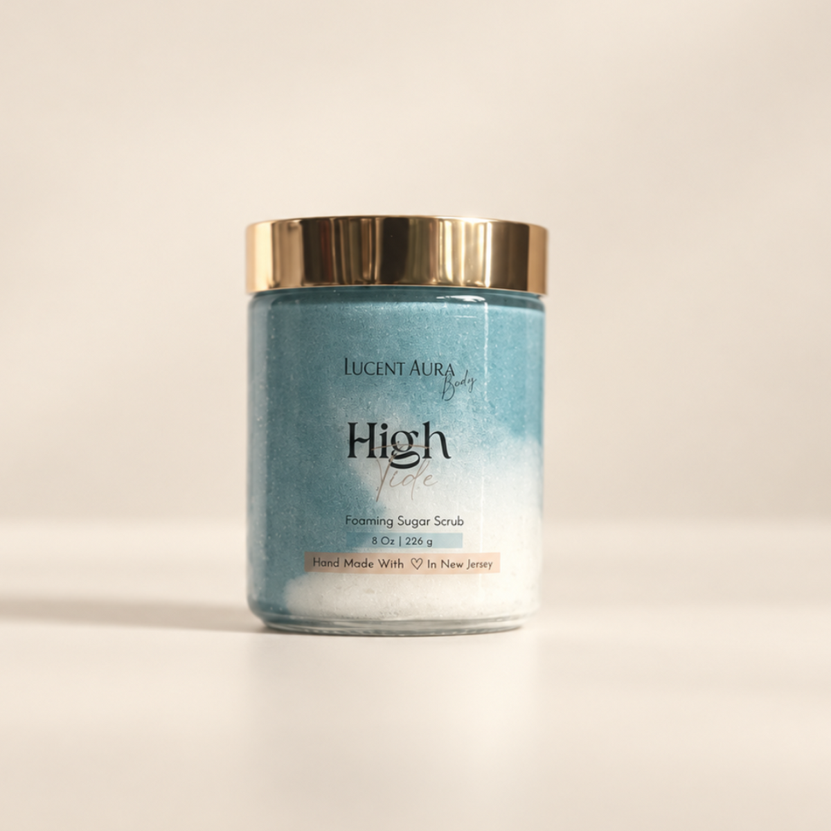 Lucint Aura High Tide Sugar Scrub in a clear glass jar with a gold metallic lid, set against a warm beige background.