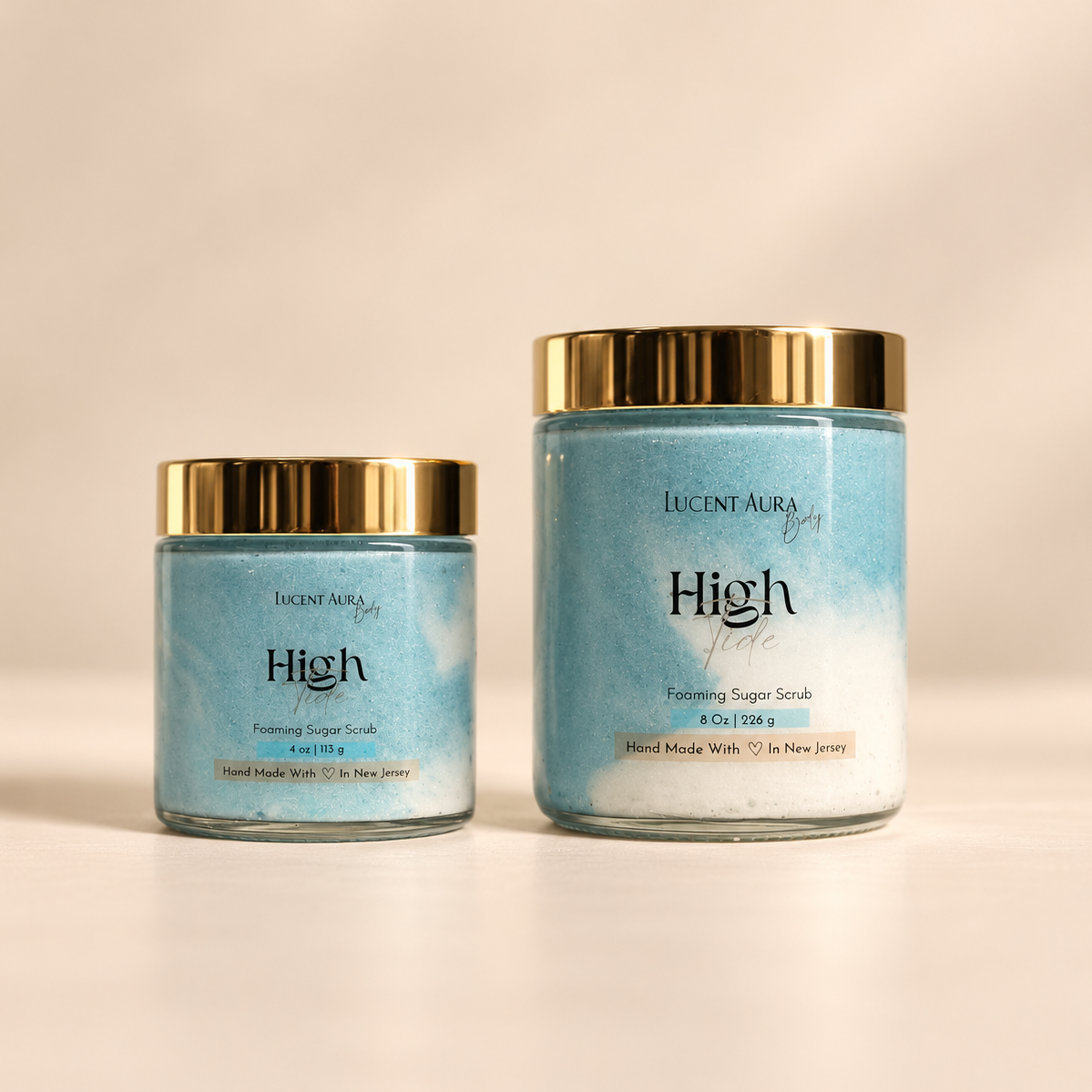 two bottles of Lucent Aura High Tide Foaming Sugar Scrub in a clear 4oz glass jar and a 8oz glass jar with a gold lids, set against a warm off-white background.