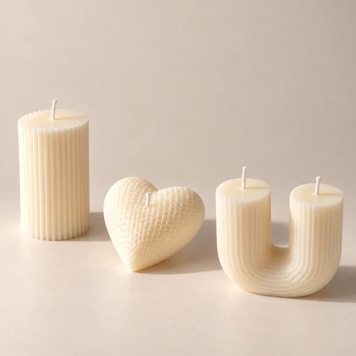 Three cream-colored decorative candles on a warm beige background: a ribbed pillar candle, a ribbed heart-shaped candle, and a ribbed U-shaped candle.