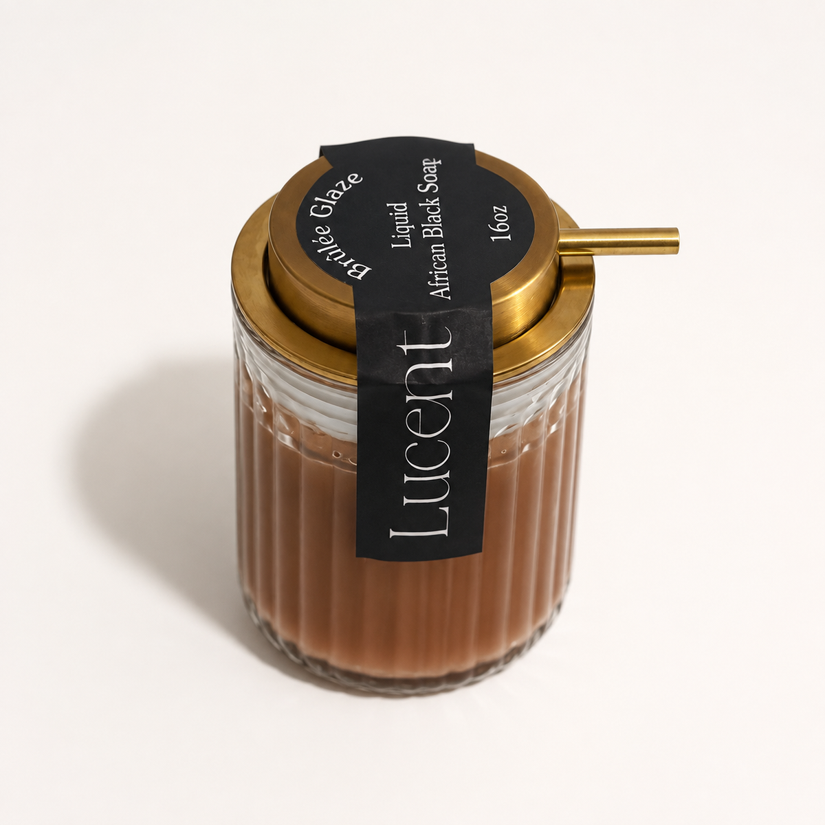 A 16oz Lucent Aura Liquid African Black Soap in a ribbed glass jar with a gold lid and a black paper label. A gold metal straw is inserted into the side of the jar, set against a clean white background.