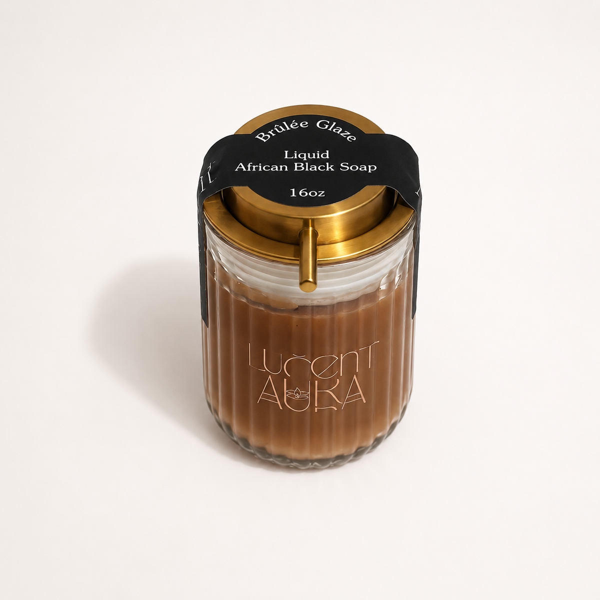 A 16oz Lucent Aura Liquid African Black Soap in a ribbed glass jar with a gold lid and a black paper label. A gold metal straw is inserted into the side of the jar, set against a clean white background.