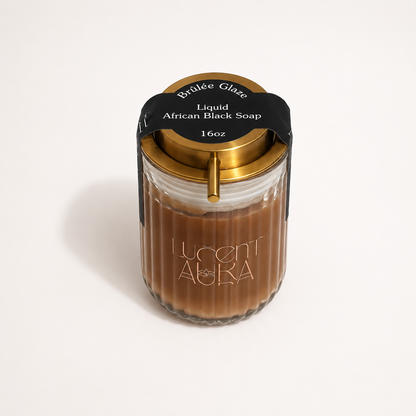 A 16oz Lucent Aura Liquid African Black Soap in a ribbed glass jar with a gold lid and a black paper label. A gold metal straw is inserted into the side of the jar, set against a clean white background.