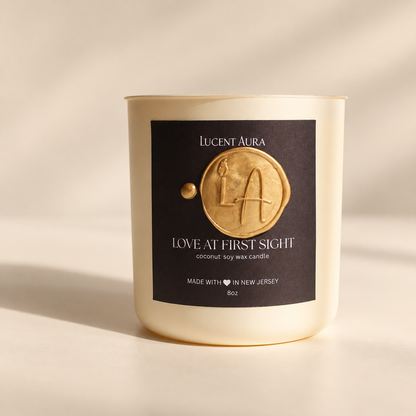 A single candle in a cream-colored glass jar with a black label and gold wax seal, photographed on a warm beige surface with soft diagonal shadows. The label reads 'Lucient Aura' and 'Love at First Sight'.