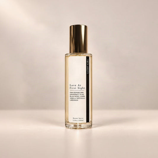 A bottle of Lucent Aura 'Love At First Sight' room spray with a gold cap and a white label, set against a warm beige background.