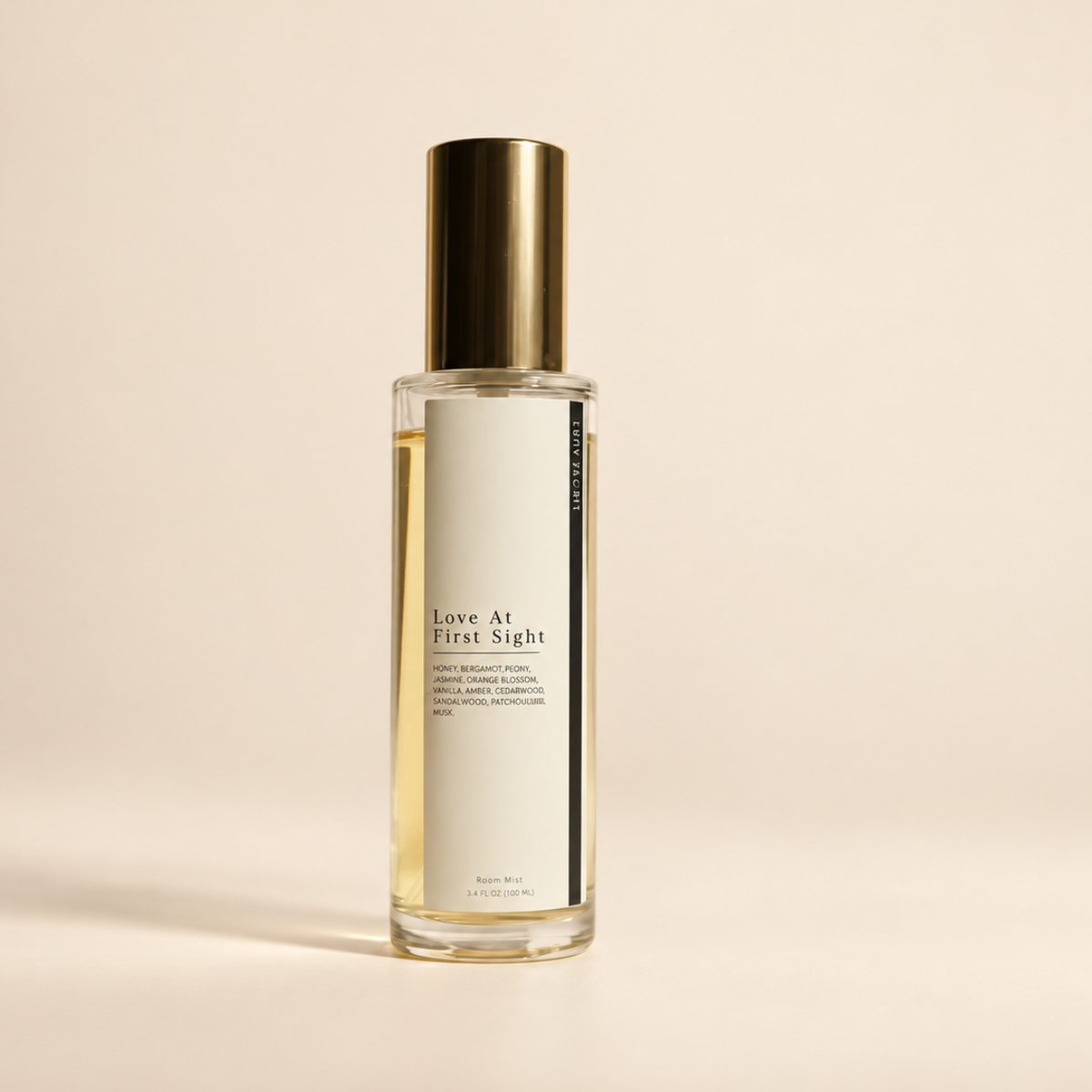 A clear glass bottle of 'Love At First Sight' room spray with a off white label and a metallic gold cap, set against a solid beige background.