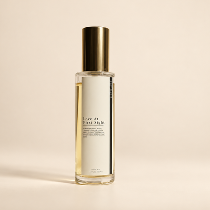 A clear glass bottle of 'Love At First Sight' room spray with a off white label and a metallic gold cap, set against a solid beige background.