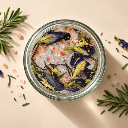 Top-down view of a clear glass jar filled with coarse bath salts and dried blue flowers, surrounded by rosemary sprigs and scattered salt crystals on a warm beige background.