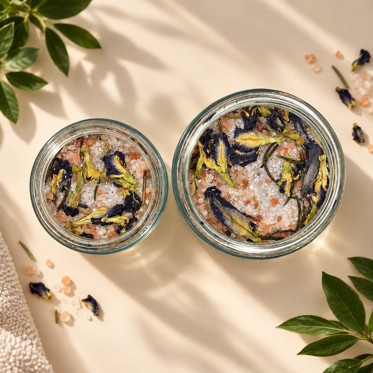 Two open clear glass jars filled with bath salts and dried botanicals, arranged on a warm beige surface with soft leaf shadows. The salts appear pink and white, mixed with dark blue and yellow flower pieces.