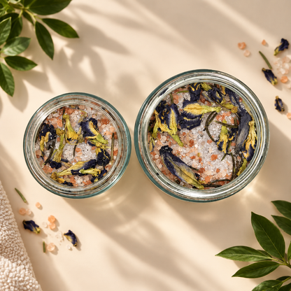 Two open clear glass jars filled with bath salts and dried botanicals, arranged on a warm beige surface with soft leaf shadows. The salts appear pink and white, mixed with dark blue and yellow flower pieces.