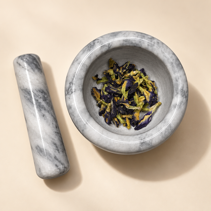 A gray and white marble mortar and pestle set on a warm beige background, with dried purple and green flowers placed inside the bowl.