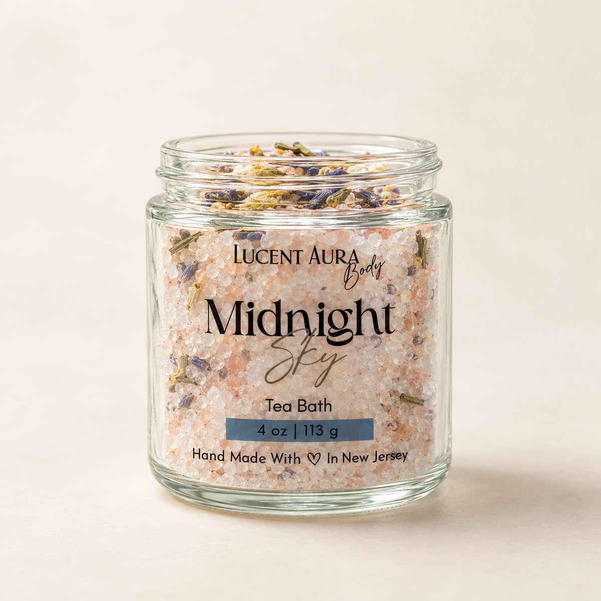 A clear glass jar filled with pink tea bath salts and dried lavender buds, labeled 'LUCENT AURA Body Midnight Sky Tea Bath' on a warm beige background.