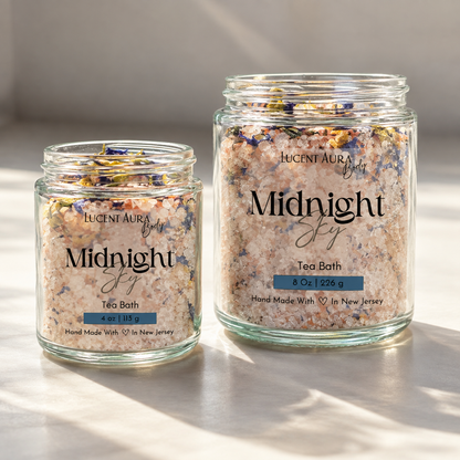 Two clear glass jars of Lucenti Aura Midnight Sky Tea Bath, one 4 oz and one 8 oz, filled with pinkish-beige bath salts and dried flower petals, set on a light surface with soft shadows.