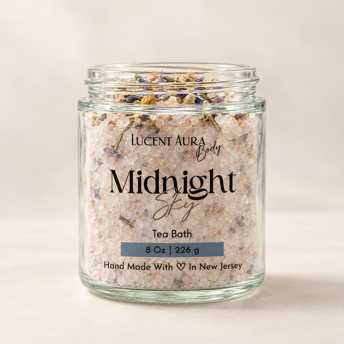 A clear glass jar of Lucent Aura Body Midnight Sky tea bath salts with dried flowers, set against a warm beige background.