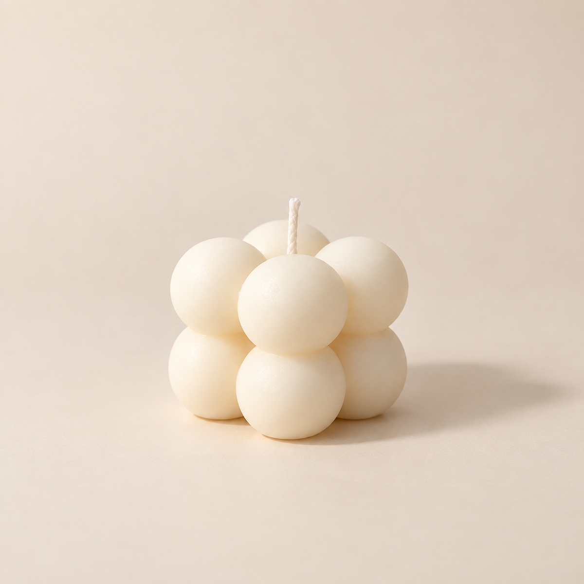 A mini bubble candle made of seven matte off-white spheres with a single wick, photographed on a warm beige studio background.