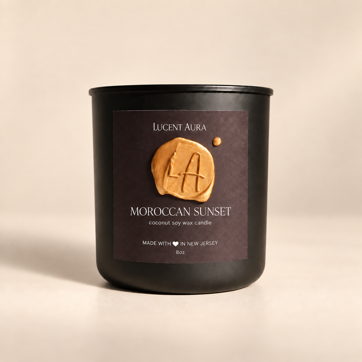 A matte black soy wax candle jar by Lucent Aura, labeled 'Moroccan Sunset', set against a warm beige background. The front label features a textured golden-brown wax seal with the brand initials 'LA'.