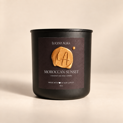 A matte black soy wax candle jar by Lucent Aura, labeled 'Moroccan Sunset', set against a warm beige background. The front label features a textured golden-brown wax seal with the brand initials 'LA'.