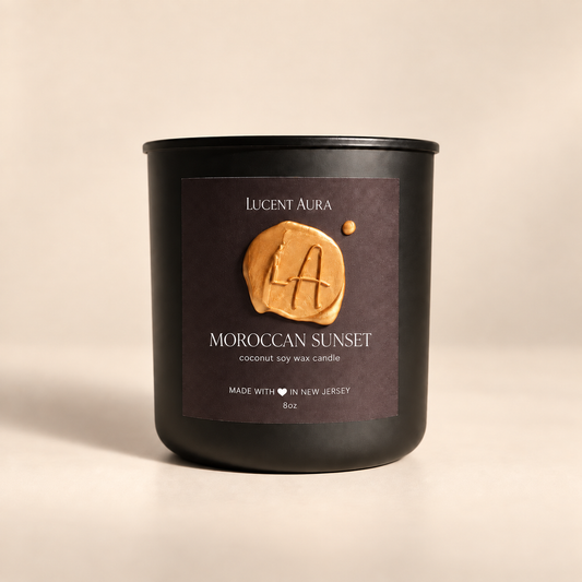 A matte black soy wax candle jar by Lucent Aura, labeled 'Moroccan Sunset', set against a warm beige background. The front label features a textured golden-brown wax seal with the brand initials 'LA'.